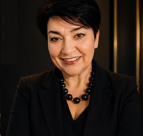 Professional headshot of a woman with short dark hair wearing a black blazer and beaded necklace, smiling against a dark background